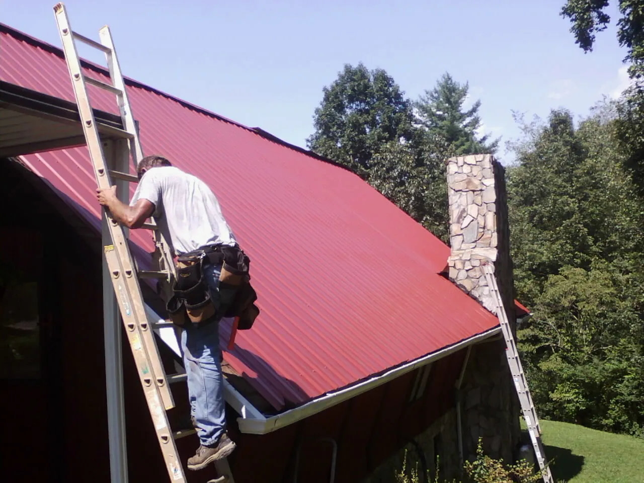 Roofer on a ladder beside a red metal roof and stone chimney — Soffit & Fascia Repair by Roofing Brothers in Zumbrota